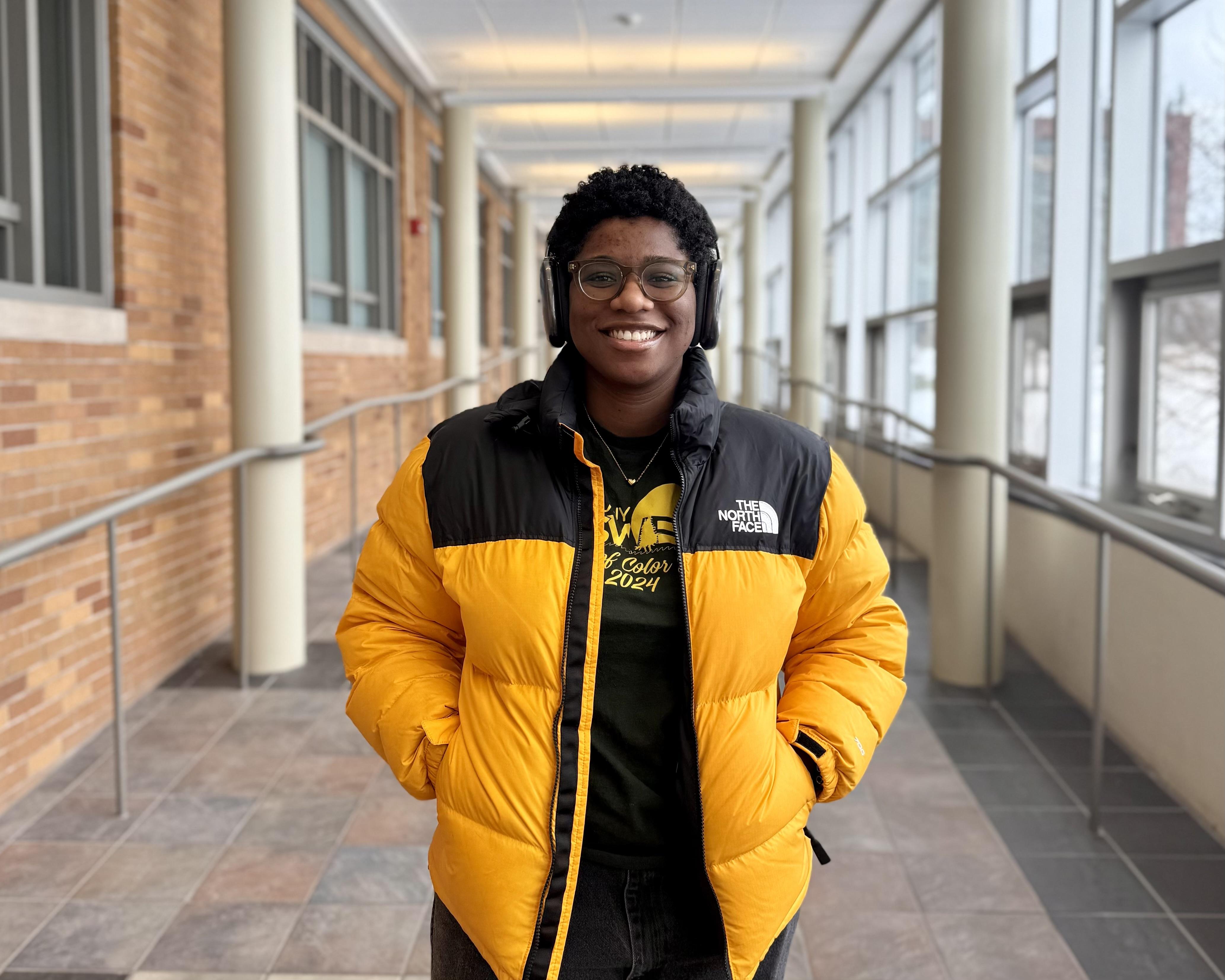 Oswego graduate student Keturah Hancock in Marano Campus Center. She wears a yellow coat and headphones.