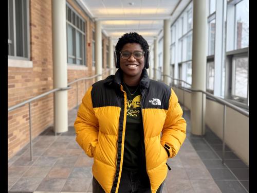 Oswego graduate student Keturah Hancock in Marano Campus Center. She wears a yellow coat and headphones.