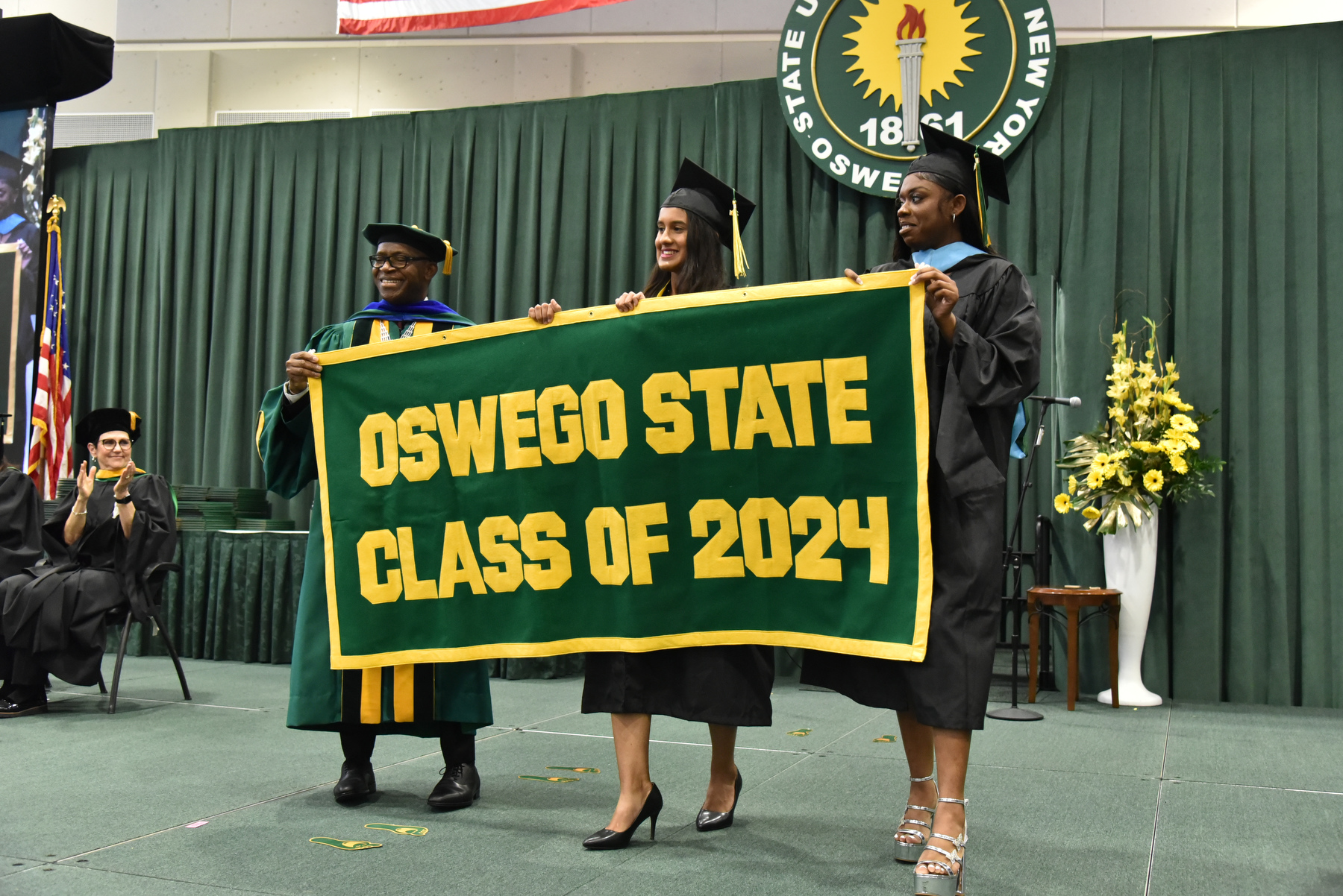 Jolie Santiago (center) and Kiara Husbands present the Class of 2024 alumni banner with President Peter O. Nwosu near the conclusion of the School of Communication, Media and the Arts and School of Education ceremony.