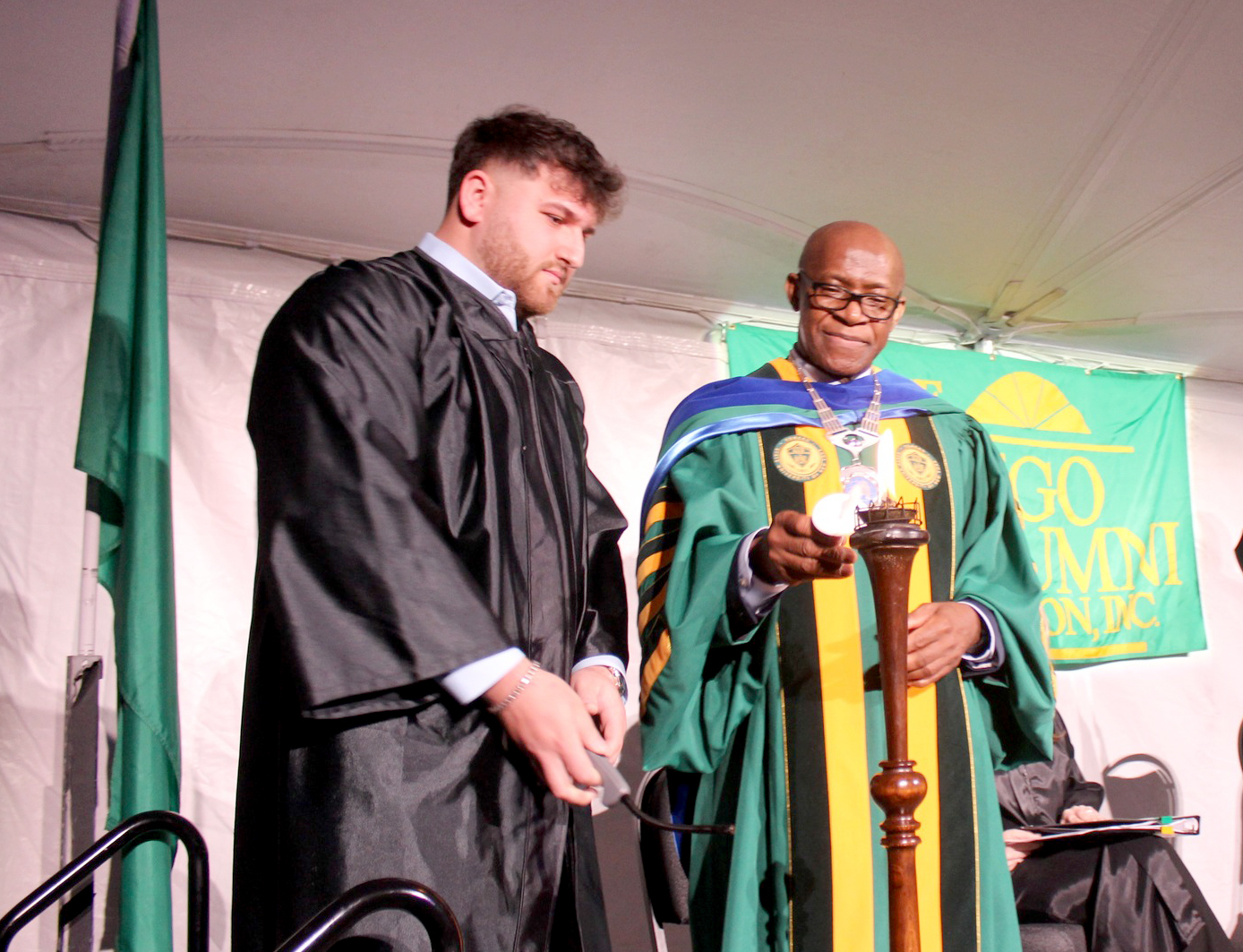 SUNY Oswego President Peter O. Nwosu lights the “flame of knowledge,” the central symbol of Commencement Eve Torchlight, before passing it to Torchbearer Matthew Bernstein of the Class of 2024, who is the son of the ceremony’s alumni emcee Brad Bernstein '89.
